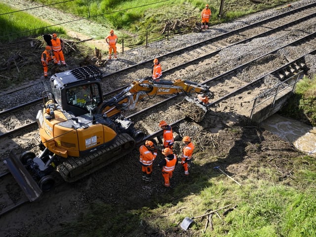 Bauarbeiter in orangefarbenen Schutzanzügen arbeiten neben Bahngleisen mit einem Bagger.