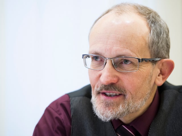 Ein Mann mit grauen Haaren, grauem Bart und schwarzer Brille.