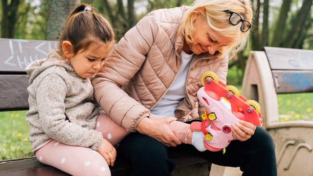 Ein Mädchen (links) und eine Frau (rechts) sitzen auf einer Bank. Die Frau zieht dem Mädchen einen Rollschuh aus.