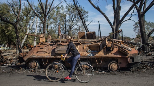 Frau fährt auf Fahrrad vor zerstörtem Panzer vorbei.