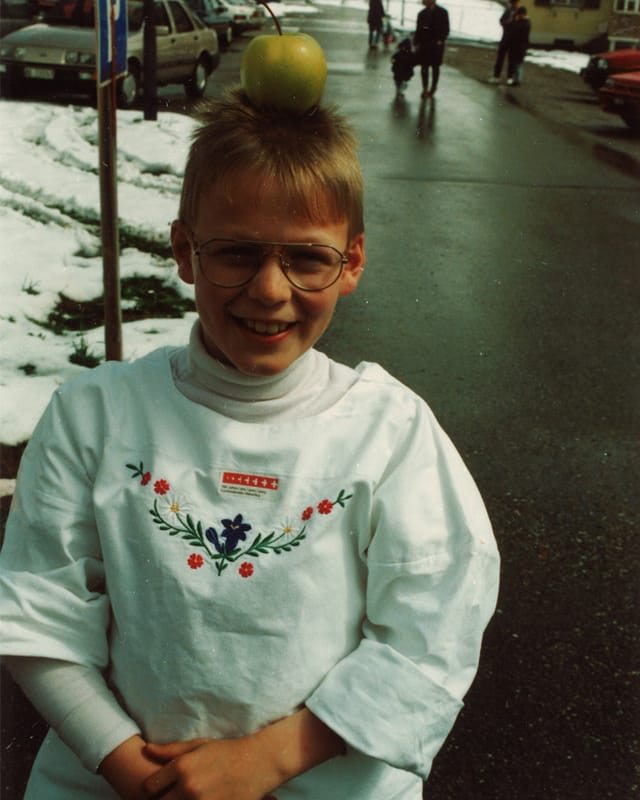 Ein blondhaariger Bub mit einem weissen bestickten Hemd trägt einen Apfel auf dem Kopf.