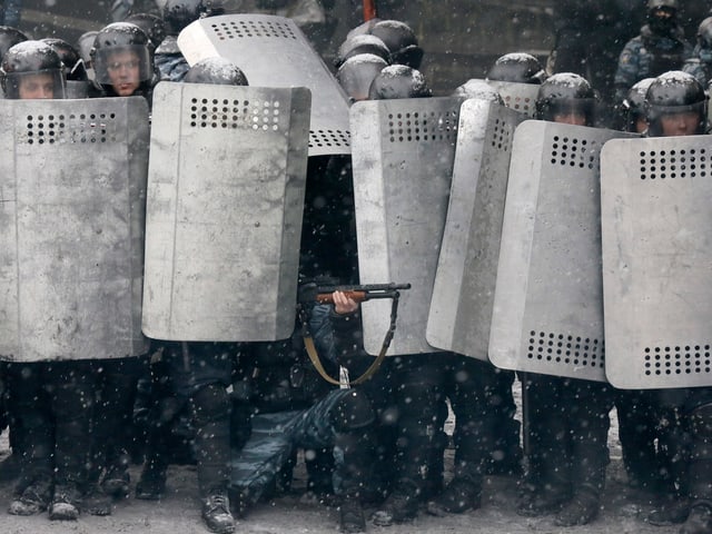 Eine Reihe von Polizisten in Vollmontur hinter ihren Schutzschildern, einer von ihnen kniet und zielt mit einem Gewehr zwischen den Schildern hindurch auf Demonstranten, die im Bild aber nicht zu sehen sinid.