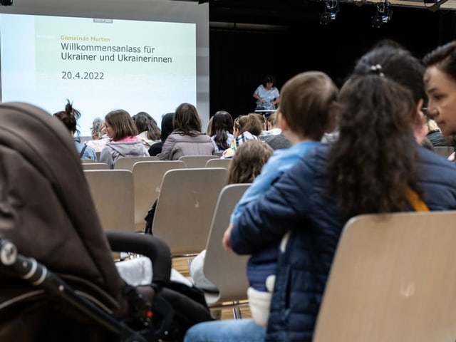 Menschen sitzen auf Stühlen. Im Hintergrund sieht man eine Leinwand. 