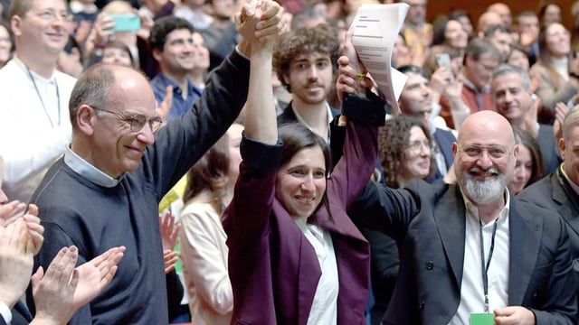 Elly Schlein bei ihrer Wahl zur Parteichefin im vergangenen März. Zwei weisse Männer halten ihre Hände hoch.