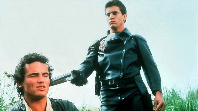 A young man in a leather suit aims a pistol at the head of a seated man.