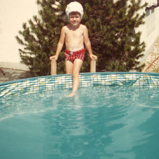 Ein kleiner Bub trägt eine Badekappe und eine Badehose und steigt über eine kleine Treppe in einen Pool.