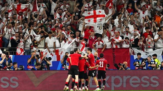 Fussballspieler jubeln auf dem Rasen, auf den Zuschauerrängen werden die Flaggen Georgiens hochgehalten.