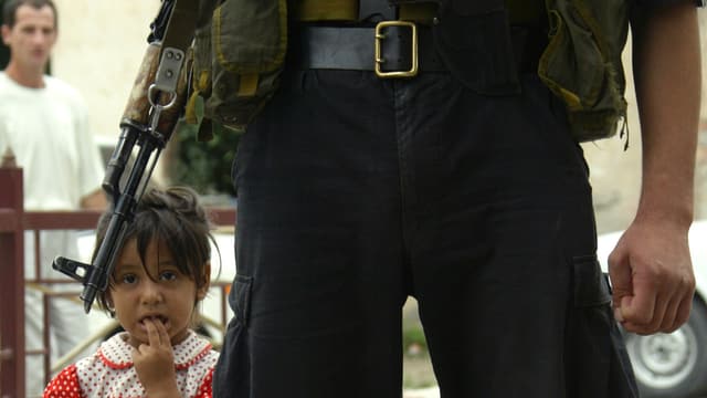 Nahhaufnahme eines tschetschenischen Soldaten (Hüfthöhe) mit Gewehr, neben ihm steht ein Mädchen.