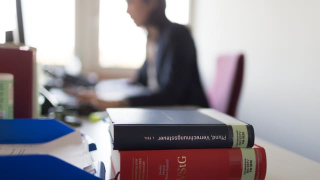 Zwei Bücher liegen auf einem Pult, im Hintergrund arbeitet eine Person.