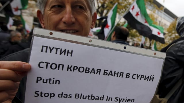 Mann hält ein A-Blatt hoch mit der Botschaft "Putin, stopp das Blutbad in Syrien" in verschiedenen Sprachen