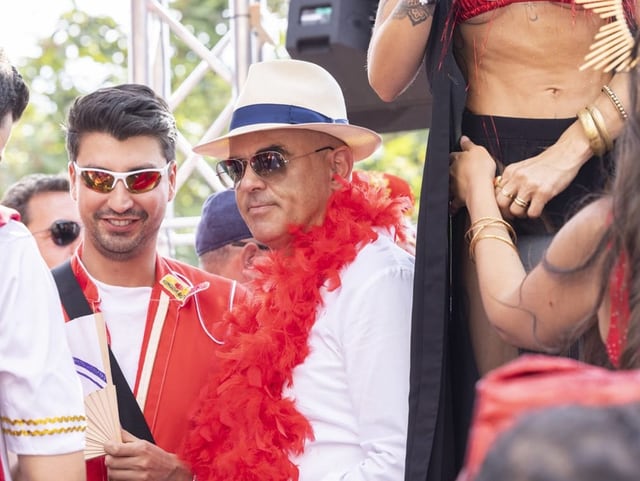 Alain Berset inmitten von Ravern mit roter Federboa, Sonnenbrille und Hut