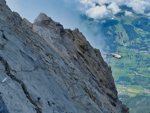 Drohne fliegt neben steilem Berghang über grünes Tal.