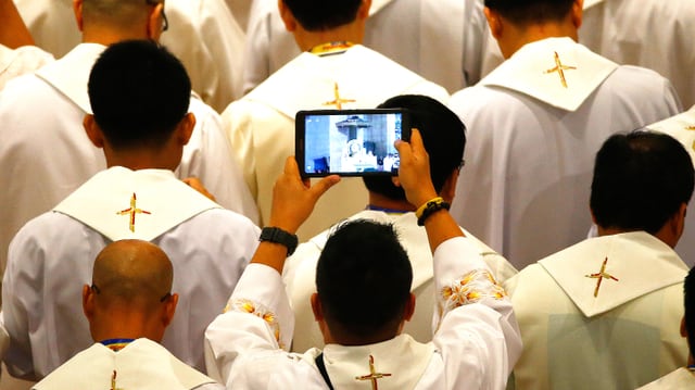 Viele Priester von hinten. Einer hält ein Tablet hoch und filmt den Papst bei seiner ersten Ansprache in Manila.