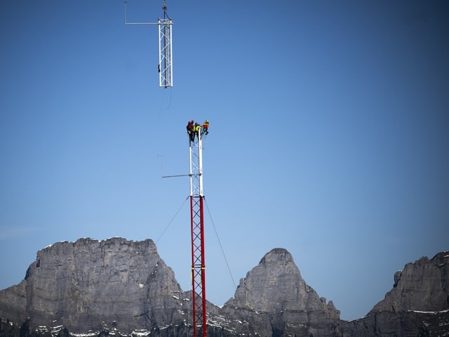 Arbeiter auf einem Funkmast vor Bergkulisse.