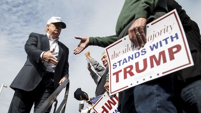 Trump mit weisser Baseballcap und Anzug schüttelt einem Anhänger die Hand, der ein Transparent hält mit der Aufschrift: «Die schweigende Mehrheit steht hinter Trump.»