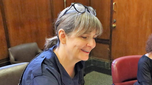 Lächelnde Frau mittleren Alters mit Brille auf dem Kopf sitzt an einem Tisch.