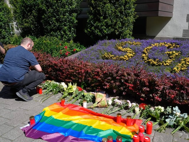 Mann kniet vor Blumen und Regenbogenflagge bei einem Mahnmal.