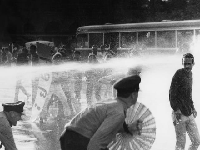 Foto von Menschen, die durch Wasserstrahlen eines Wasserwerfers gehen, während ein Bus im Hintergrund steht.