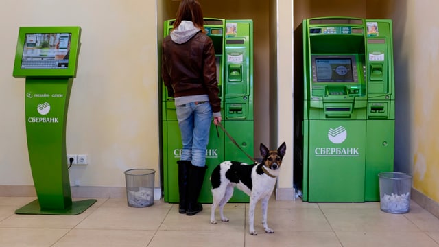 Eine Frau mit einem Hund an einer Leine bezieht Geld an einem grünen Bankomaten.