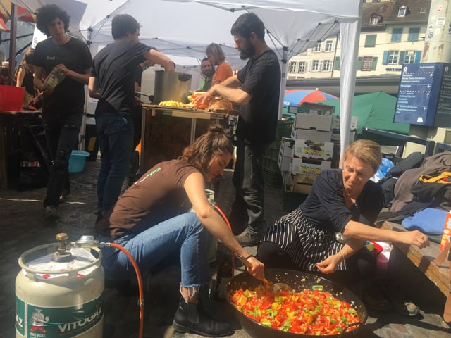 vorne kochen zwei Frauen auf einem improvisierten herd, hinten rüsten Männer Gemüse.