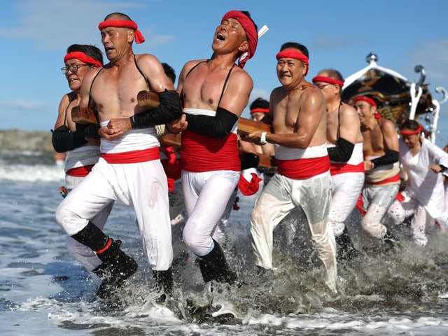 Gruppe von Männern in traditioneller Kleidung zieht einen Festwagen durch das Wasser.