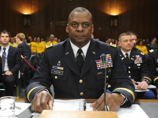 Lloyd Austin is sitting at a table in a room with a large crowd. 