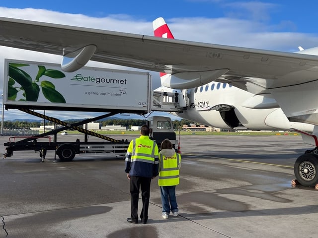 Zwei Menschen mit gelben Leuchtwesten stehen unter dem Flügel eines Flugzeuges. 