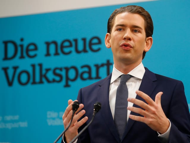 Kurz gestikuliert vor einem Parteiplakat.