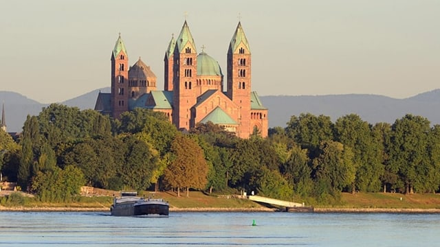 Ein grosser Dom mit rötlicher Fassade und grünen Dächern. Umgeben von Bäumen an einem Fluss. Im Hintergrund Hügel.