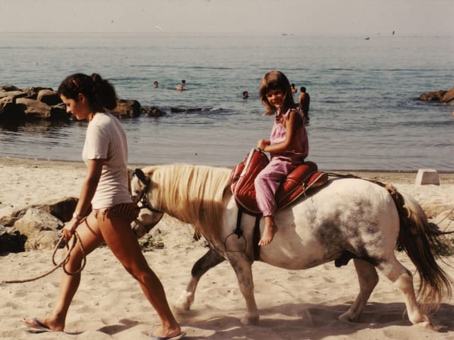 Eine Frau führt ein Pony, auf dem ein kleines Mädchen sitzt, am Zaum an einem Strand entlang.