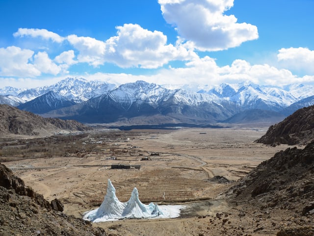 Zwei Eiskegel in brauner Landschaft im Vordergrund. Dann ein Dorf. Im Hintergrund hohe, schneebedeckte Berge.