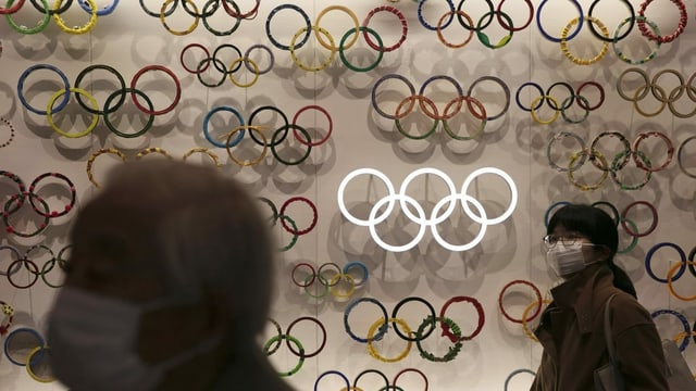 People in masks walk past a wall with Olympic rings.