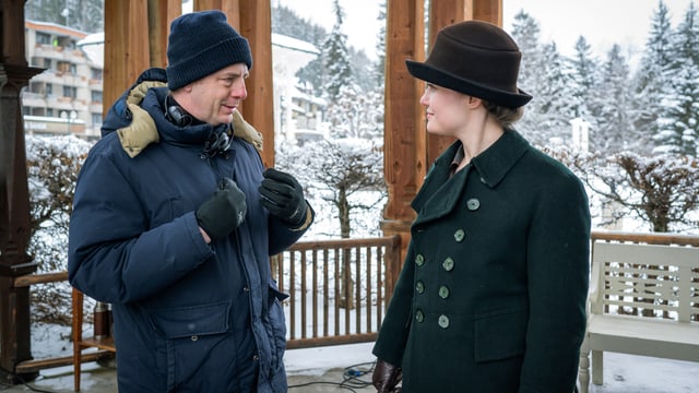 Mann im Winteranorak und Frau in historischem Kostüm stehen sich am Filmset gegenüber und sprechen miteinander.