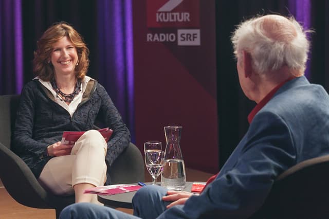 Eva Oertle unterhält sich mit Franz Hohler, der von hinten zu sehen ist. Sie lächelt.