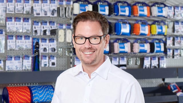 Der Mann mit Brille heisst Marcel Meister. Im Hintergrund sind Garne und Schnüre. Er steht in einem Laden.