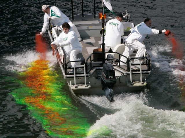 Personen kippen ein oranges Pulver in den Fluss, der sich dann grün verfärbt.