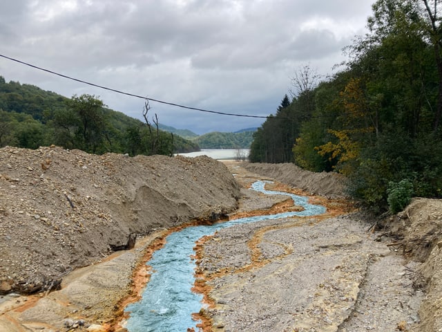 Ein grell-hellblauer Bach fliesst durch ein braunes Flussbett in einen grauen See.