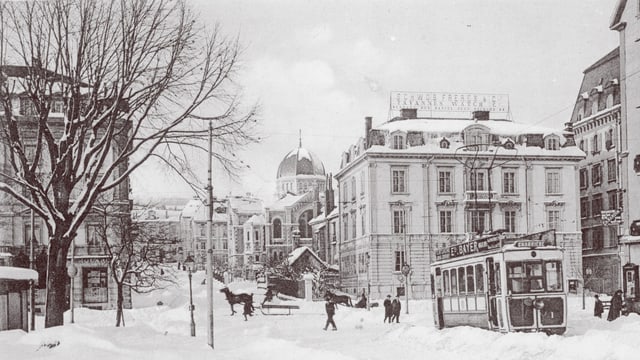 Blick auf eine verschneite Strasse, gesäumt von herrschaftlichen Häusern, darauf unterwegs ein Tram und eine Kutsche.