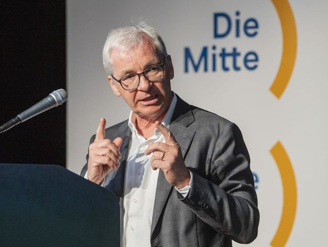 Ein Mann mit weissem Haar und Brille vor dem Logo «Die Mitte».