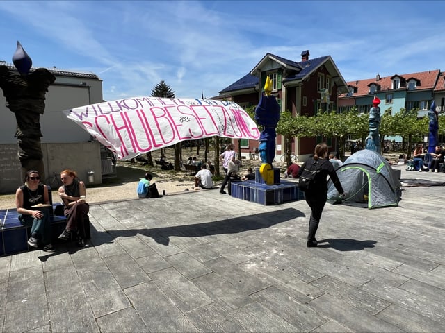 Menschen auf einem Platz mit einem beschrifteten Banner und Zelten.