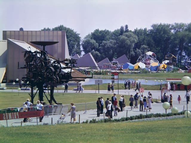 Expo 64 Lausanne: Menschenmenge im Freien mit Skulpturen und geometrischen Strukturen im Hintergrund.