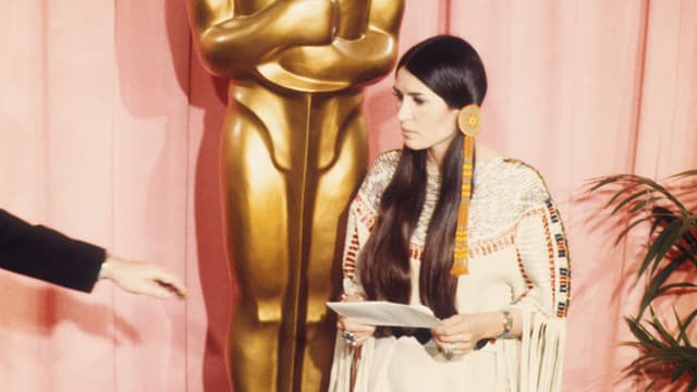 Aktivistin Sacheen Littlefeather steht in traditioneller indianischer Kleidung vor einer grossen Oscar-Statue.