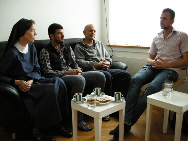 Eine Nonne und drei Männer sitzen auf einer Couch und einem Stuhl um einen Kaffeetisch.