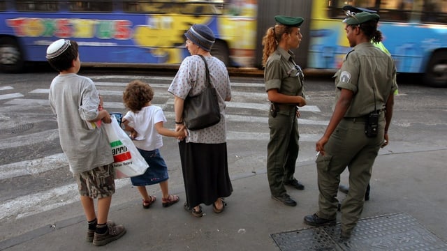 Orthodoxe Jüdin mit zwei Kindern mit Kippa am Strassenrand vor einer Krezung, daneben drei Polizisten, davon eine Polizistin.