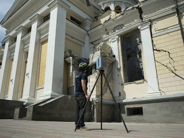 Eine gelb-weisse Fassade mit Rissen, davor ein Mann mit Helm, vor ihm ein Gerät mit Stativ.