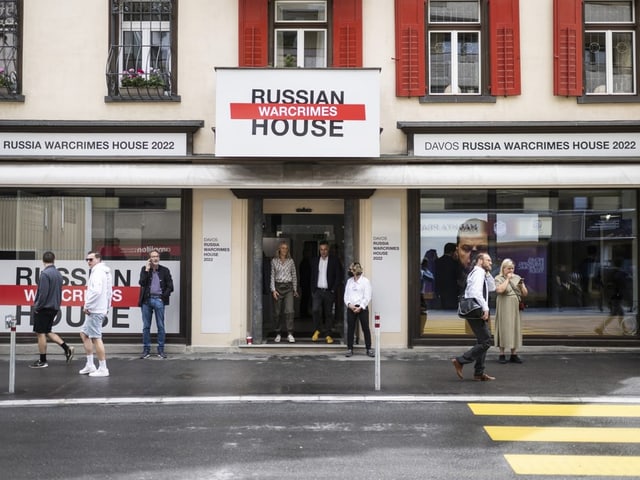 Blick auf ein Haus an einer Strasse. Es ist beschriftet mit «Russian Warcrimes House. Menschen laufen davor.