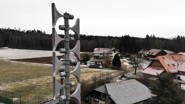 Foto einer grauen Sirene, total acht Sirenen übereinander, dahinter Häuser und schneebedeckte Wiese.