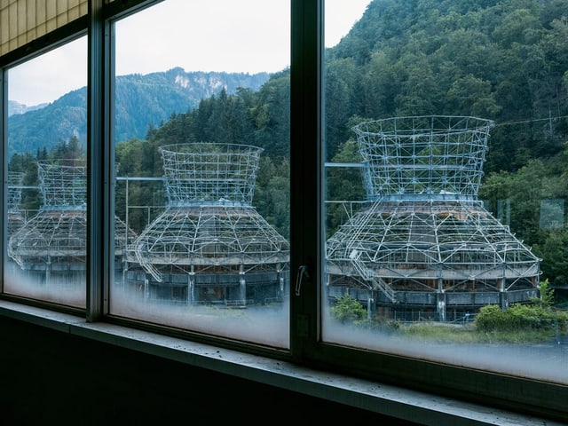 Blick aus einem Fenster auf industrielle Strukturen vor einem bewaldeten Hügel.