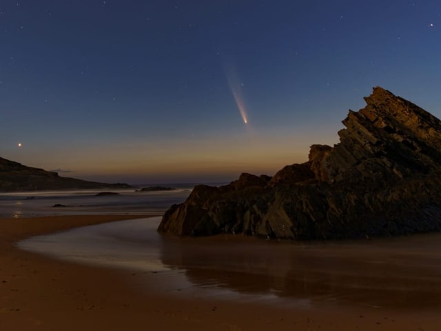 Strand bei Nacht mit Komet am Himmel und Felsen im Vordergrund.