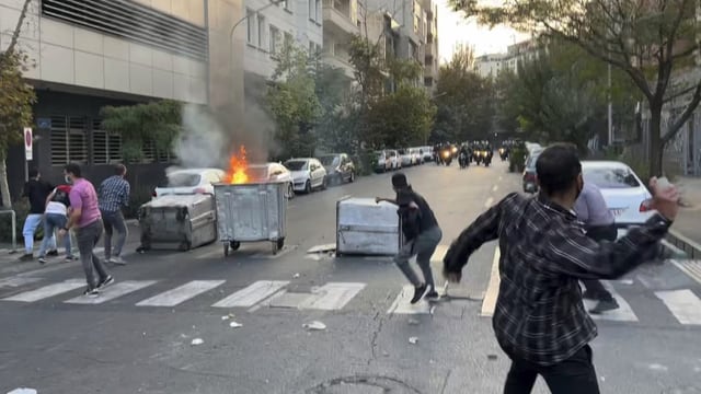 Junge Männer werfen Steine auf Polizisten auf Motorrädern. Brennender Abfallcontainer.